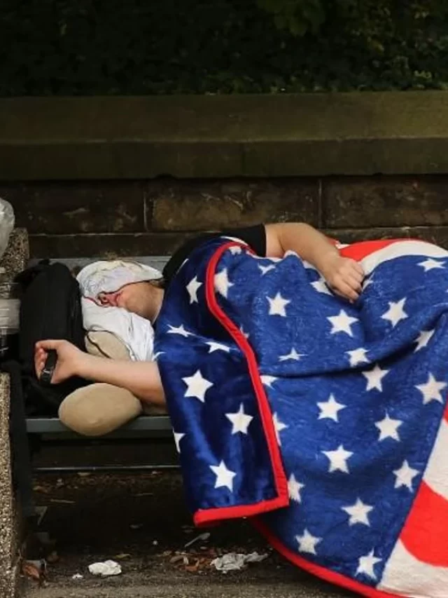cropped-a-homeless-man-sleeps-under-an-american-flag-blanket-on-a-park-bench-on-september-10-2013-in-the-brooklyn-borough-of-new-york-c.webp